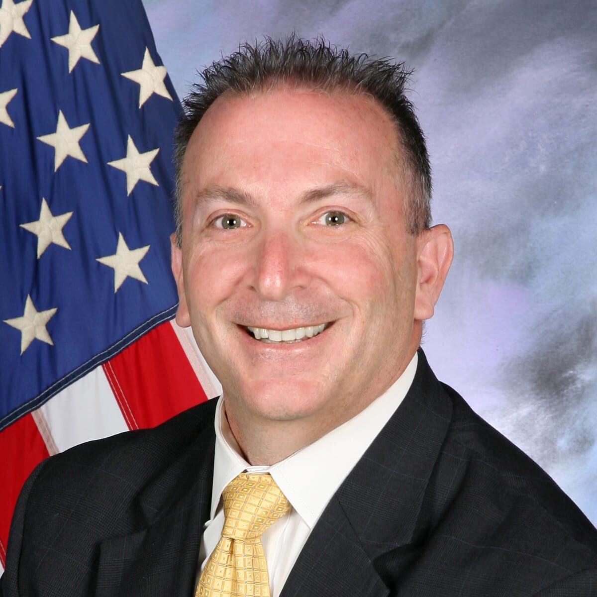 Support staff Richard Koplar, smiling man wearing suit with yellow tie in front of American flag