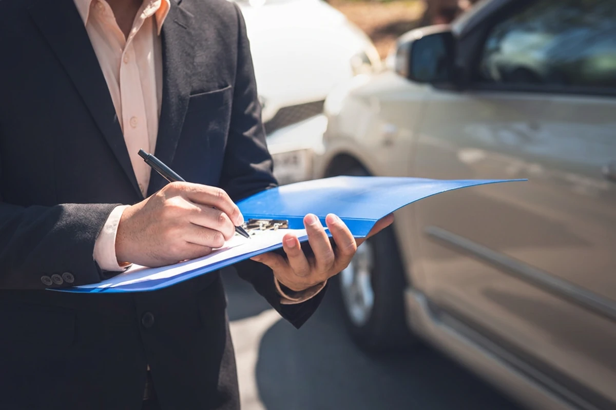 A man in a black suit with a light-colored shirt writes on a document on a blue clipboard. Blurry damaged cars are visible in the background, indicating an accident.
